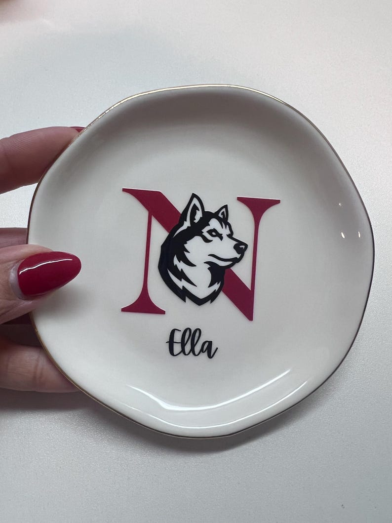 Puede incluir: Platito de cer&aacute;mica blanca con borde dorado. Presenta la letra "N" en granate con un gr&aacute;fico de perro husky en blanco y negro. El nombre "Ella" est&aacute; escrito en cursiva negra debajo.