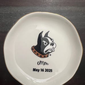 Puede incluir: Un plato de cer&aacute;mica blanco con borde dorado. El plato presenta un gr&aacute;fico de Boston Terrier en blanco y negro con un collar marr&oacute;n. El texto "cmm" y "May 16 2025" est&aacute;n impresos debajo.