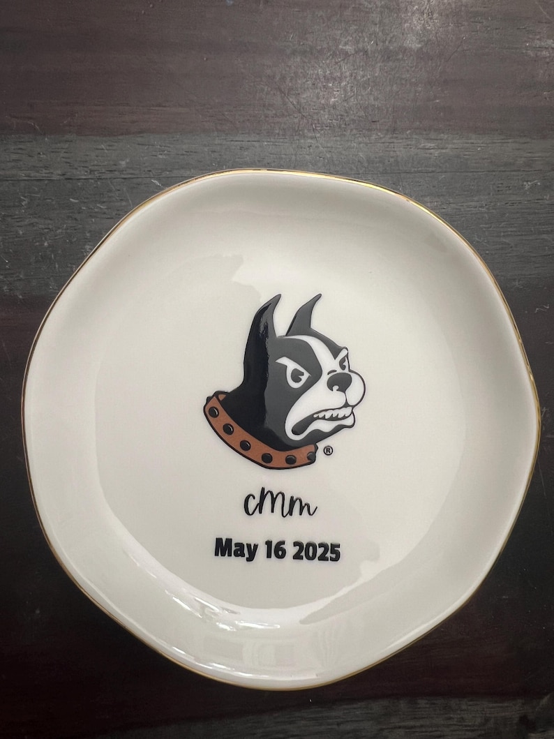 Puede incluir: Un plato de cer&aacute;mica blanco con borde dorado. El plato presenta un gr&aacute;fico de Boston Terrier en blanco y negro con un collar marr&oacute;n. El texto "cmm" y "May 16 2025" est&aacute;n impresos debajo.