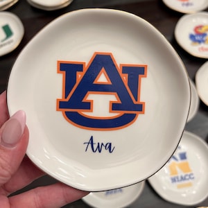 Puede incluir: Platito de cer&aacute;mica blanca con borde dorado, con el logo de la Universidad de Auburn en azul y naranja. El nombre "Ava" est&aacute; escrito debajo del logo. Otros platos similares est&aacute;n en el fondo.