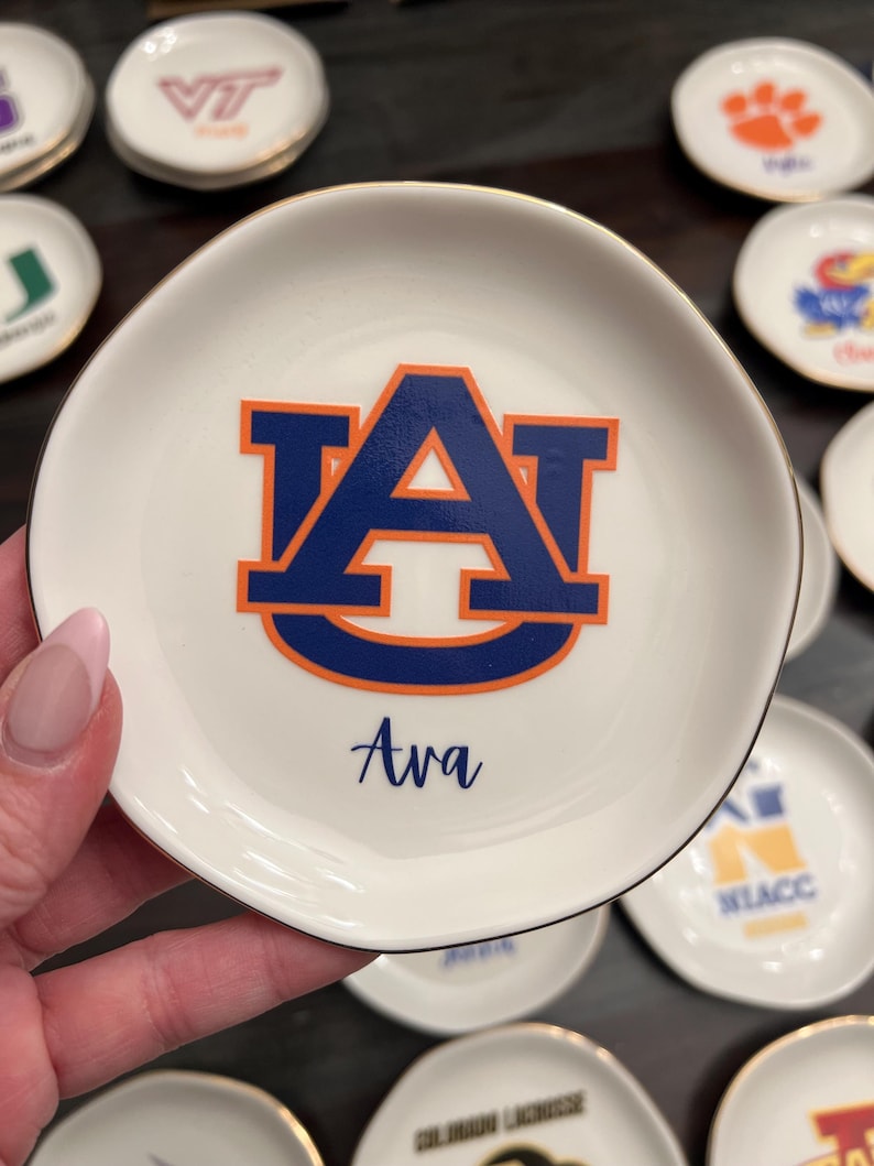 Puede incluir: Platito de cer&aacute;mica blanca con borde dorado, con el logo de la Universidad de Auburn en azul y naranja. El nombre "Ava" est&aacute; escrito debajo del logo. Otros platos similares est&aacute;n en el fondo.