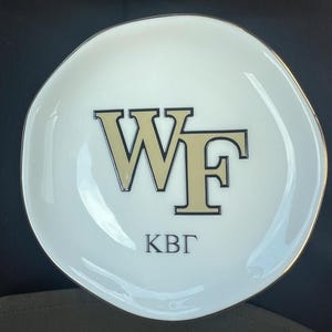 Puede incluir: Un plato de cer&aacute;mica blanco con borde dorado. El plato presenta las letras "WF" en dorado con contorno negro, y las letras "KB&Gamma;" en negro debajo. El plato tiene una forma irregular, ligeramente ondulada.