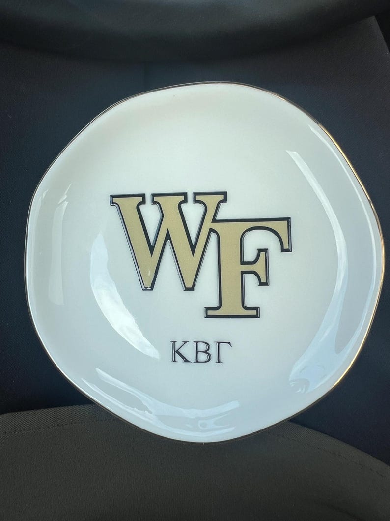 Puede incluir: Un plato de cer&aacute;mica blanco con borde dorado. El plato presenta las letras "WF" en dorado con contorno negro, y las letras "KB&Gamma;" en negro debajo. El plato tiene una forma irregular, ligeramente ondulada.