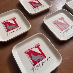 Puede incluir: Platos cuadrados blancos con bordes dorados. Cada plato presenta un logotipo 'N' rojo con un lobo gris, las palabras "WOMEN'S LACROSSE" y "COUNT ON ME", adem&aacute;s de un nombre y un n&uacute;mero en escritura negra. Los platos est&aacute;n colocados sobre una superficie marr&oacute;n.