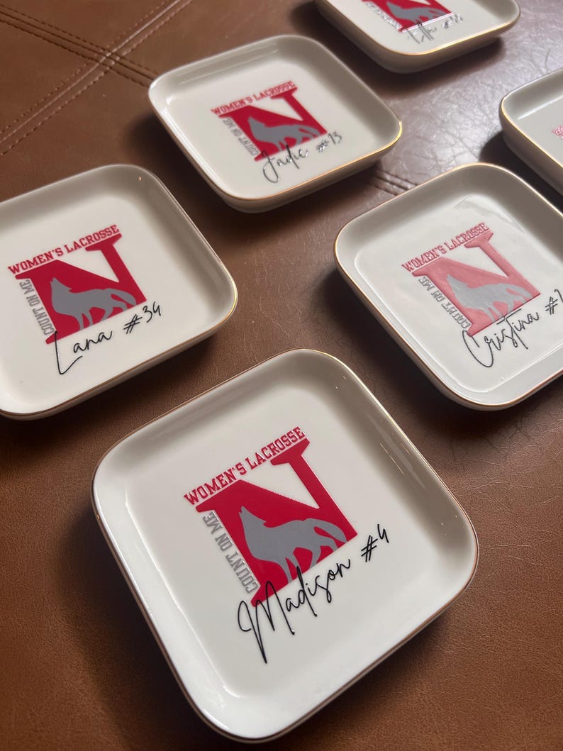 Puede incluir: Platos cuadrados blancos con bordes dorados. Cada plato presenta un logotipo 'N' rojo con un lobo gris, las palabras "WOMEN'S LACROSSE" y "COUNT ON ME", adem&aacute;s de un nombre y un n&uacute;mero en escritura negra. Los platos est&aacute;n colocados sobre una superficie marr&oacute;n.