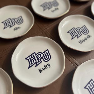 Puede incluir: Una colecci&oacute;n de peque&ntilde;os platos de cer&aacute;mica blanca con borde dorado. Cada plato presenta las letras azul marino "HPU" y un nombre en fuente cursiva. Los platos est&aacute;n dispuestos sobre una superficie marr&oacute;n.