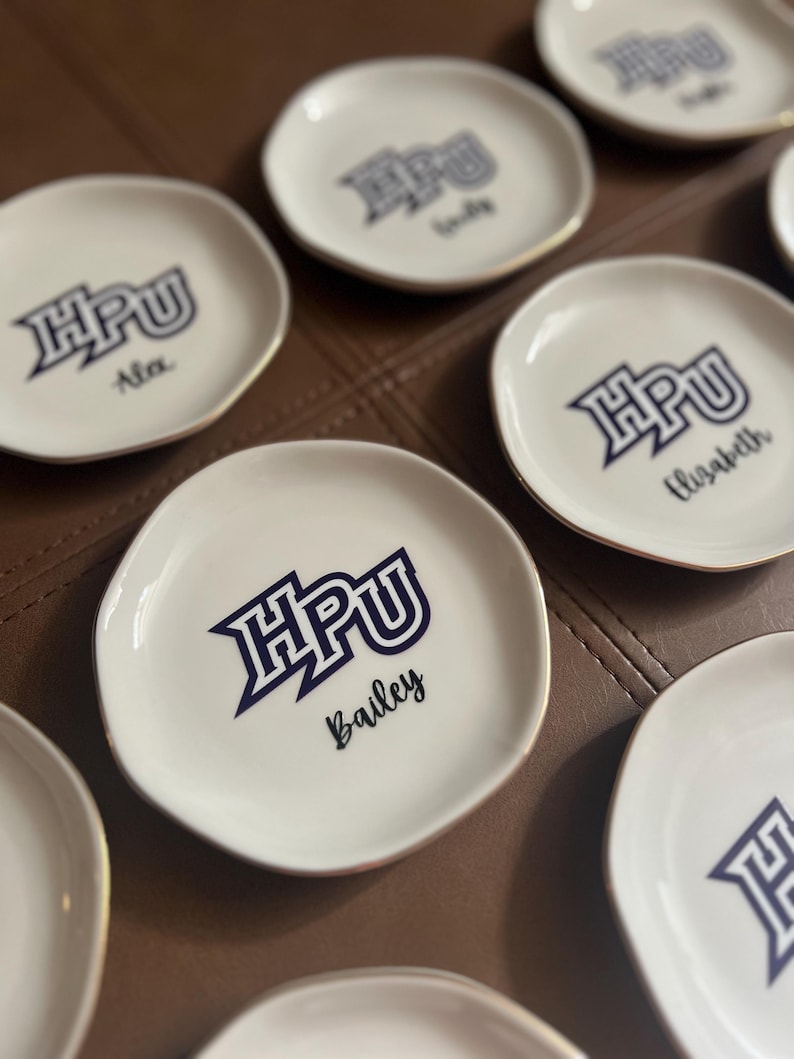 Puede incluir: Una colecci&oacute;n de peque&ntilde;os platos de cer&aacute;mica blanca con borde dorado. Cada plato presenta las letras azul marino "HPU" y un nombre en fuente cursiva. Los platos est&aacute;n dispuestos sobre una superficie marr&oacute;n.