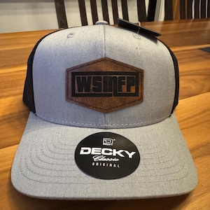 May include: A gray and black trucker hat featuring a brown hexagonal leather patch on the front with the black text WSMFP. The hat has a flat brim and a circular black sticker on the brim that reads DECKY Classic ORIGINAL in white text.