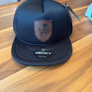 Puede incluir: Una gorra tipo trucker negra con un parche de cuero marr&oacute;n en forma de escudo que muestra un gr&aacute;fico negro de una figura corriendo. La gorra tiene una visera plana con una pegatina negra que dice DECKY ORIGINAL y un cord&oacute;n negro al frente.