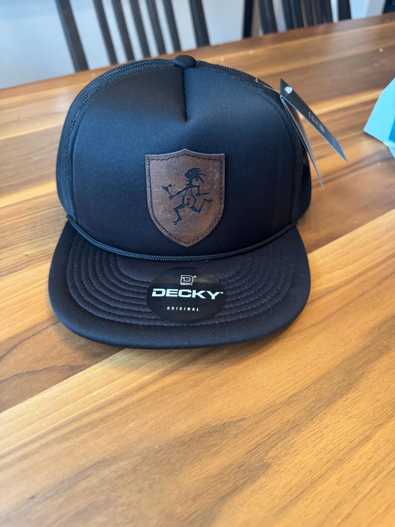 Puede incluir: Una gorra tipo trucker negra con un parche de cuero marr&oacute;n en forma de escudo que muestra un gr&aacute;fico negro de una figura corriendo. La gorra tiene una visera plana con una pegatina negra que dice DECKY ORIGINAL y un cord&oacute;n negro al frente.