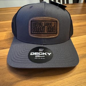 Può includere: Un cappellino da baseball grigio antracite con retro in rete nera e visiera piatta. Una toppa rettangolare marrone è centrata sulla parte anteriore con il testo "NEVER MISS A SUNDAY SHOW" in nero. Un adesivo nero circolare sulla visiera riporta "DECKY Classic ORIGINAL".