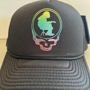 May include: Black trucker hat with a rainbow-colored graphic of a person sitting on a stool inside a skull. The skull has a pink outline.