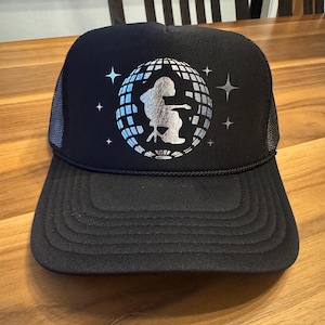 May include: A black trucker hat featuring a silver metallic graphic of a disco ball with a silhouette of a person playing a guitar inside. The design is surrounded by small four-pointed star accents. The hat has a structured front and mesh back.