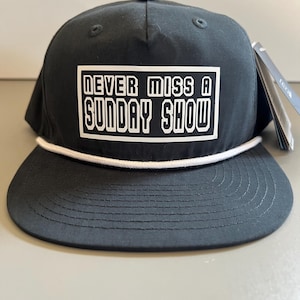 Könnte beinhalten: Eine schwarze Baseballkappe mit einem weißen rechteckigen Aufnäher auf der Vorderseite, der den Text "NEVER MISS A SUNDAY SHOW" in einer fetten, weißen serifenlosen Schrift zeigt. Die Kappe hat eine weiße Kordel am Kronenansatz und einen flachen Schirm.