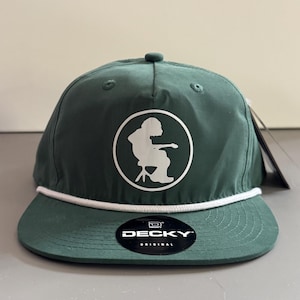Peut inclure: Casquette de baseball vert foncé avec un détail de corde blanche et une visière plate. Le chapeau présente un graphisme circulaire blanc d'une personne assise sur une chaise. La marque "DECKY" est affichée sur un autocollant noir.