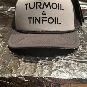 Könnte beinhalten: Eine weiße und graue Trucker-Kappe mit schwarzem Text, der "TURMOIL & TINFOIL" lautet.