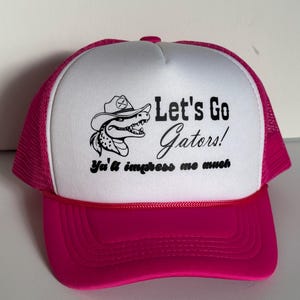 May include: A pink and white trucker hat with the text "Let's Go Gators!" and a cartoon alligator wearing a cowboy hat. The hat has a pink brim and mesh back.