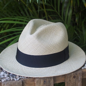 Op de afbeelding: Een lichtbeige strohoed met een zwarte lintband. De hoed ligt op een houten oppervlak met kleine stenen.