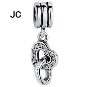 May include: Silver charm featuring two intertwined hearts, one of which is embellished with small, clear crystals. The charm is designed to be attached to a bracelet or necklace. The letters "JC" are visible in the upper left.