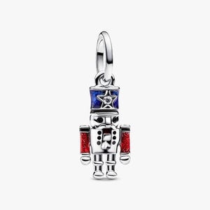 May include: A silver-coloured charm depicting a nutcracker soldier. The charm features a blue and red colour scheme, with a star on the blue hat. The arms are red with glitter. It has a loop at the top for attaching to a bracelet or necklace.