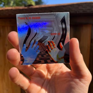 May include: A square holographic sticker featuring a first-person perspective of a hallway with a geometric carpet pattern. The text "There is no escape" and "wake up" is printed in red. Two hands are visible in the foreground, one blue and one orange.