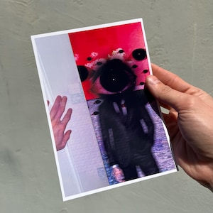 May include: A rectangular art print featuring a surreal illustration of a figure in a black suit with a large, singular eye for a head. The background transitions from a vibrant red top section with floating black orbs to a purple textured base.