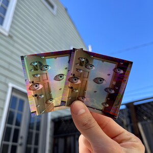 May include: Two iridescent stickers with a holographic finish depicting a door with multiple eyes. The stickers are held in a hand against a green house background.