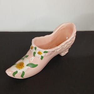 May include: A pink ceramic shoe planter with white flowers and green leaves painted on it.