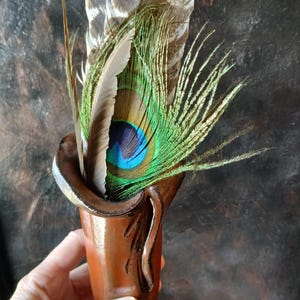 May include: A brown ceramic vase with a flared rim, filled with a variety of feathers. The feathers include large brown and white patterned ones, green and brown feathers, and a vibrant peacock feather with blue and green details.