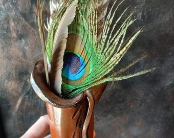 May include: A brown ceramic vase with a flared rim, filled with a variety of feathers. The feathers include large brown and white patterned ones, green and brown feathers, and a vibrant peacock feather with blue and green details.