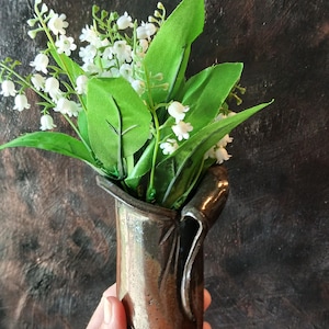 Może przedstawiać: Brązowy ceramiczny wazon z bukietem sztucznych konwalii. Kwiaty mają małe, białe kwiaty w kształcie dzwonka i zielone liście. Wazon ma unikalny, teksturowany design.