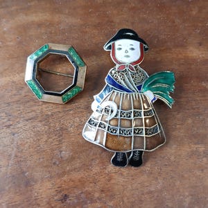 May include: Two enamel brooches, one is a green and gold octagon with a black center, the other is a girl in a brown and white plaid dress with a green hat and a green and white bouquet.