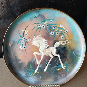 May include: A round decorative plate with a metallic rim featuring a central white horse in motion. The background is a blend of blue, pink, and dark tones with painted white flowers and green foliage accents surrounding the horse figure.