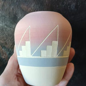 Peut inclure: Un vase en céramique peint à la main avec une palette de couleurs pastel. Le vase présente un dégradé rose clair et bleu, avec un motif géométrique blanc. La base est de couleur crème, et une bande bleu clair sépare la base du corps principal.