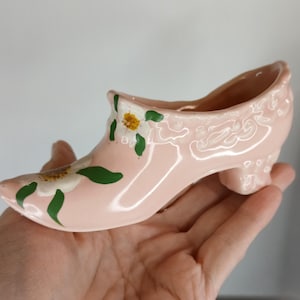 May include: A pink ceramic shoe planter with white flowers and green leaves painted on it. The shoe has a high heel and a pointed toe.