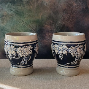 Op de afbeelding: Twee keramische bekers met een blauw en wit druivenrankenontwerp. De bekers hebben een witte glazuur en een blauwe basis met witte druivenranken en druiven.
