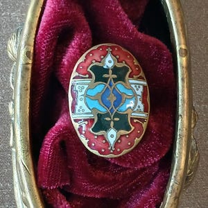May include: An oval-shaped, ornate brooch with a detailed design in red, green, blue, and white enamel. The brooch is set in a gold-colored metal frame and rests on a bed of deep red velvet.