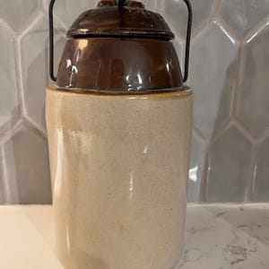May include: A cylindrical stoneware crock featuring a light beige body and a dark brown glazed lid. The container includes a dark metal wire handle that arches over the top, secured by a central metal latch mechanism on the lid's surface.