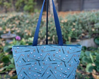 May include: A light blue tote bag with a golf-themed pattern featuring golf clubs and golf balls. The bag has two exterior pockets and navy blue handles. The bag is made of fabric and is designed for carrying items.