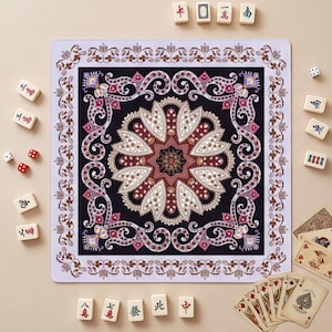 Puede incluir: Un tapete de juego cuadrado con un diseño de mandala central y patrones de cachemira intrincados en tonos negro, blanco, burdeos y dorado. El borde tiene un motivo floral repetitivo. Fichas de mahjong y cartas de juego rodean el tapete.