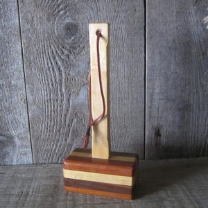 May include: A wooden gavel with a rectangular base composed of horizontal stripes in shades of brown and tan. A light-colored wooden handle extends upwards, with a leather strap attached at the top. The gavel is set against a rustic wooden background.