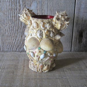 May include: A cylindrical vase completely covered in a variety of beige, tan, and white seashells of different shapes and sizes. The top rim of the vase features two prominent spiky shells on either side, and the interior is a deep red color.
