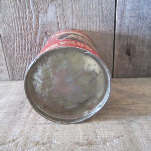 K&ouml;nnte beinhalten: Vintage Bierdose aus Metall mit rotem und goldenem Etikett. Der Boden der Dose ist sichtbar und weist Gebrauchsspuren auf. Die Dose steht auf einer verwitterten Holzoberfl&auml;che, was auf eine rustikale &Auml;sthetik hindeutet.