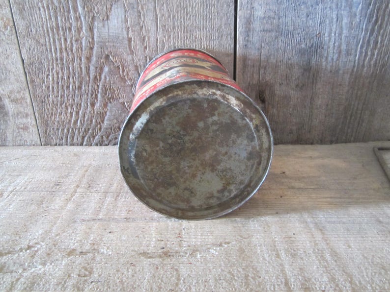 K&ouml;nnte beinhalten: Vintage Metallkanne mit rostigem Boden und rot-gold gestreiftem Deckel. Die zylindrische Dose weist Alters- und Abnutzungserscheinungen auf, was darauf hindeutet, dass es sich um ein antikes oder sammelw&uuml;rdiges Objekt handelt. Die Dose steht vor einem Holzgrund.