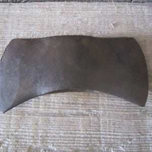 May include: A double-bit axe head made of dark, weathered metal. The object features two symmetrical, curved cutting edges on opposite sides and a central eye for a handle. The surface shows signs of oxidation and age with a textured, matte finish.