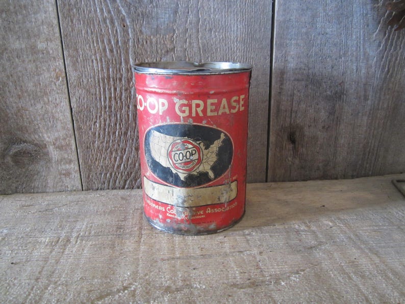 K&ouml;nnte beinhalten: Rote, vintage Metall-Dose mit der Aufschrift "CO-OP GREASE" und einer Karte der Vereinigten Staaten. Die Dose weist Gebrauchsspuren auf. Der Text "CO-OP GREASE" ist wei&szlig;. Die zylindrische Dose steht auf einer Holzoberfl&auml;che. Antikes Design.