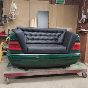 Op de afbeelding: Een unieke groene bank geconstrueerd uit de achterkant van een auto, met een zwart, met knopen bekleed leren zitvlak. De bank heeft rode achterlichten en staat op een platform met wielen, wat een mix van auto- en meubeldesign laat zien.