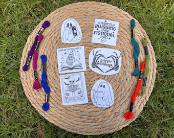 May include: A collection of white stickers with black illustrations and text, including ghosts, skeletons, and open books. The text includes "SPREAD THOSE PAGES", "Trigger warnings", and "CURRENTLY BLUSHING OVER FICTIONAL men + ". Embroidery floss in various colors surrounds the stickers.