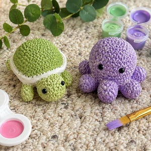 May include: A light green crocheted turtle and a light purple crocheted octopus with black plastic eyes. The set includes a white plastic paint palette with three colors, a blue paintbrush, and several small open containers of colorful acrylic paint.