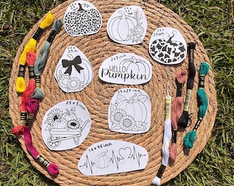 May include: A collection of black and white pumpkin-themed designs, some with floral accents, and the text "Hello Pumpkin." The designs are on white paper, with measurements in inches. Embroidery floss in various colors surrounds the designs.